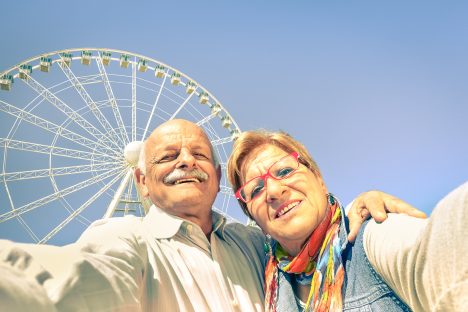 Happy retired senior couple taking selfie at Rimini beach - Concept of active playful elderly with mobile phone - Mature people fun lifestyle in sunny day with strong sunlight color tones - Photo taken in public area edited with vintage filtered look