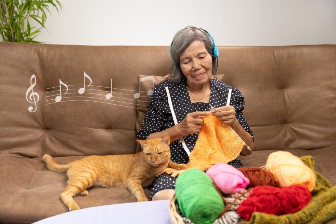 Older woman sitting on the couch with a cat by her side, knitting while listening to music through headphones.