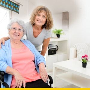 Woman leaning over an elderly lady in a wheelchair with her arm over her, both smiling and facing the camera in the living room