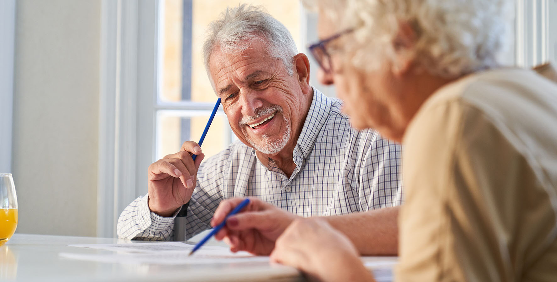 An older man and older lady look at paperwork. They have pencils in their hand. The lady is out of focus and the man is the main focus of the image. the man is smiling. They are in a home setting.