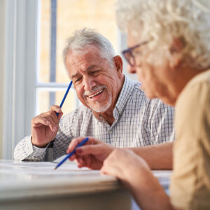 An older man and older lady look at paperwork. They have pencils in their hand. The lady is out of focus and the man is the main focus of the image. the man is smiling. They are in a home setting.